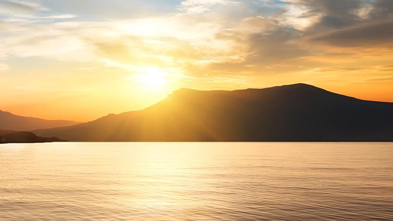 The sun setting behind a mountain overlooking calm waters